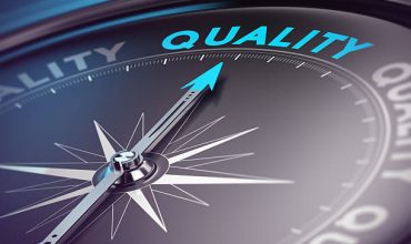 Compass needle pointing the blue text. Blue and black tones with blur effect and focus on the main word. Concept for quality assurance management.